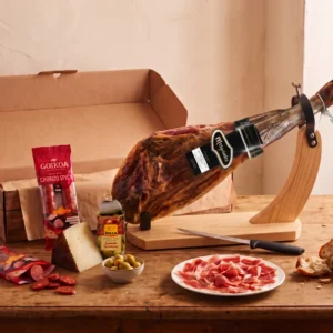 Whole Melquiades 50% Ibérico ham with its black brand label on a ham stand, with Goikoa chorizo, Manchego wedge and a Jolca tin of olives stuffed with Serrano ham