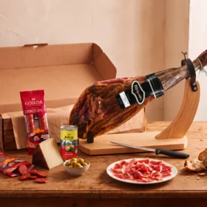 Whole Melquiades 50% Iberico ham with its black brand label on a ham stand, with Goikoa chorizo, Manchego wedge and a Jolca tin of olives stuffed with Serrano ham