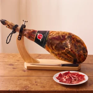 Whole Cortijo de Canata Serrano ham with its black brand label on a pine ham stand, with a ham knife and a plate of sliced jamón